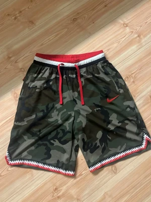 Nike camo shorts med röda detaljer - Säljer ett par camoflagemönstrade shorts från Nike med röda och vita detaljer. Shortsen har elastisk midja med röd snörning och en röd Nike-logga på benet. Snygg zigzag-detalj längs bensluten. Perfekta för en sportig look.