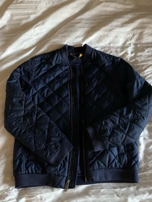 Mörkblå Ralph Laurent jacka - Snygg mörkblå Ralph Laurent jacka med quiltat mönster och ribbade muddar. Storlek 14-16 Jackan har dragkedja framtill, två sidofickor med knapp och en klassisk bomberkrage. Materialet är glansigt och ger en modern look. Perfekt för dig som vill ha en stilren och trendig jacka.