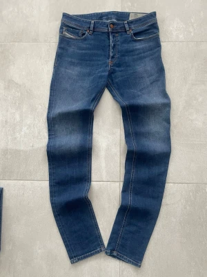 Blå slim fit jeans från Diesel - Snygga blå slim fit jeans från Diesel med klassisk femficksmodell och bruna kontrastsömmar. Jeansen har en skinnpatch med Diesel-logga bak i midjan och är tillverkade i mjukt denimtyg. Perfekta för en avslappnad och trendig look.