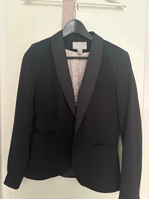 Svart kavaj från H&M, storlek 38 - Stilren svart kavaj från H&M med klassisk krage och två fickor framtill. Fodret är ljusrosa med svarta prickar, vilket ger en cool kontrast. Perfekt för dig som vill ha en enkel men snygg look. Materialet är mjukt och bekvämt.