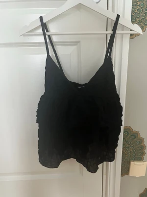 Svart volang linnetopp - Säljer en svart linnetopp med tunna axelband och volangdetaljer. Toppen har en luftig och lätt känsla, perfekt för varma dagar eller som ett lagerplagg. Enkel att matcha med både jeans och kjol.