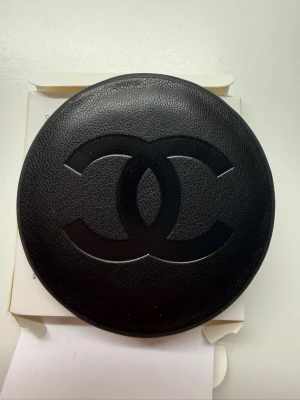 Chanel rund sminkspegel i svart läder - Snygg rund sminkspegel från Chanel med svart läderfodral och den klassiska Chanel-loggan på framsidan. Spegeln är kompakt och perfekt att ha i väskan för snabba touch-ups. Lyxig känsla och stilren design som verkligen sticker ut.