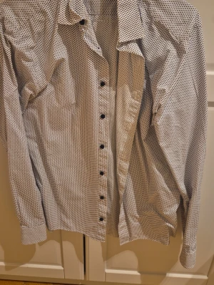 Vit skjorta med blå prickar The Shirt Factory - Snygg vit skjorta med små blå prickar och svarta knappar. Tillverkad i 100% bomull vilket gör den skön och luftig. Klassisk krage och långärmad modell, perfekt för en clean och stilren look.