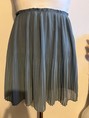 Plisserad blå kortkjol - Säljer en snygg plisserad kortkjol i en mjuk blå nyans. Kjolen har elastisk midja och ett lätt, luftigt tyg som ger ett fint fall. Perfekt för dig som gillar en enkel men stilren look. Färgen är smutsig blå. Finns små skvanker 