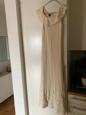 Lång beige spetsklänning med volang - Superfin långklänning i beige med spetsdetaljer och volang vid axlar och nederkant. Klänningen har ett romantiskt mönster och är perfekt för sommarens alla tillfällen. Snyggt fall och offshoulder-modell för en bohemisk look.