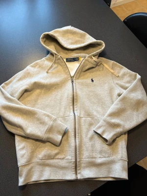 Beige hoodie från Polo Ralph Lauren - Snygg beige hoodie från Polo Ralph Lauren med dragkedja framtill, tröjan är sprillans ny, den passade bara inte min storlek.Tillverkad i mjukt bomullsmaterial som känns riktigt skönt mot huden. Perfekt för en chill och stilren look. Hoodien passar S, tveka inte på att kontakta mig vid minsta lilla
