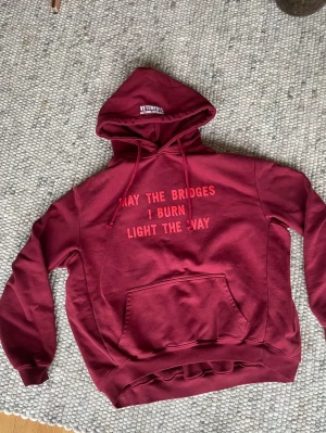 Vinröd hoodie från Vetements - Säljer en vinröd hoodie från Vetements med trycket 'MAY THE BRIDGES I BURN LIGHT THE WAY' på bröstet. Hoodien har huva med dragsko, magficka och ribbade muddar. Tillverkad i mjuk bomull för maximal komfort och streetwear-vibe.