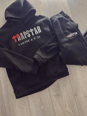 Svart Trapstar hoodie med tryck - Säljer en svart hoodie från Trapstar med stort tryck på bröstet där det står 'Trapstar It's a Secret' i vitt och rött. Klassisk huva och magficka, mjukt material som känns skönt mot huden. Perfekt för dig som gillar streetwear och vill sticka ut.
