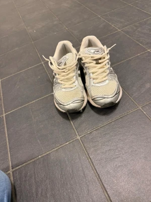 Beige och silver Asics sneakers - Säljer ett par snygga Asics sneakers i beige och silver med mesh- och syntetpaneler. Skorna har klassisk låg profil, svarta detaljer och bekväm dämpad sula. Perfekta för dig som gillar sportig stil och vill ha något som funkar till det mesta.