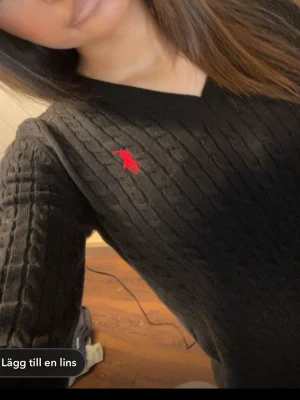 Svart kabelstickad tröja Polo Ralph Lauren - Kabelstickad svart tröja från Polo Ralph Lauren med röd broderad logga på bröstet. V-ringad hals och långa ärmar. Klassisk och tidlös design som passar till mycket. Ribbstickade muddar vid ärmslut och nederkant.