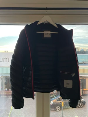 Svart dunjacka från Moncler - Snygg svart dunjacka från Moncler med quiltad design och röd dragkedja. Jackan har en innerficka med dragkedja och Moncler-logga på insidan. Perfekt för kalla vinterdagar och har en klassisk, stilren look. Dragkedjan har dock en liten defekt som ni ser på en av bilderna 