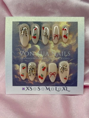 BowCore (XS);Medium Almond - Handgjorda press-on naglar i en medium almondform. Designen har en mjölkig bas med söta röda körsbär och detaljerade svarta rosetter. Perfekt för dig som vill ha en lekfull och unik look på naglarna.