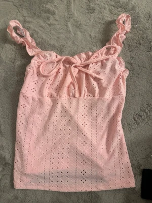 Ljusrosa broderad linnetopp - Söt ljusrosa linnetopp med broderade detaljer och volangiga axelband. Toppen har knyt framtill och är ärmlös, perfekt för varma dagar. Materialet känns lätt och luftigt, vilket gör den superbekväm att bära.