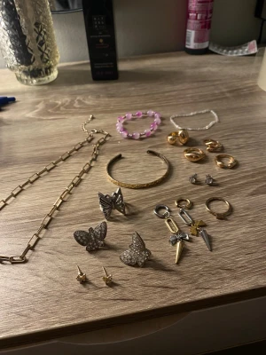 Mixat smyckespaket silver & guld - Säljer ett smyckespaket med flera accessoarer: guldigt kedjehalsband, armband i guld och rosa pärlor, örhängen i fjärilsform med stenar, chunky guldringar, små örhängen, samt coola örhängen med rosetter och spikar. Materialen är metall, pärlor och strass. Perfekt för dig som gillar att mixa stilar.