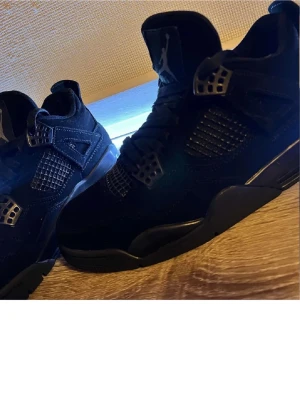 Svarta Air Jordan 4 Retro sneakers - Säljer ett par helsvarta Air Jordan 4 Retro sneakers med meshpaneler och detaljer i plast. Skorna har den klassiska Jumpman-loggan på plösen och hälen. Ovandelen är i mocka och sulan är svart. Riktigt clean och tidlös modell för dig som gillar streetwear.