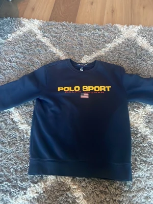 Mörkblå sweatshirt Polo Sport - Mörkblå sweatshirt från Polo Sport Ralph Lauren med gul logga och amerikansk flagga på bröstet. Klassisk rund halsringning och långa ärmar. Perfekt för en avslappnad och sportig stil.