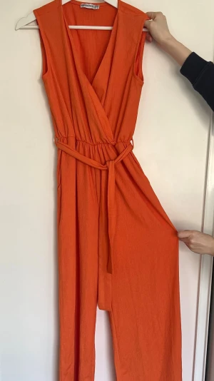Orange ärmlös jumpsuit med skärp - Snygg orange ärmlös jumpsuit  med omlott-ringning och avtagbart midjeskärp. Tillverkad i ett mjukt, lättviktigt tyg med struktur. Vida ben och resår i midjan för en bekväm passform. Perfekt för dig som vill sticka ut med färg.