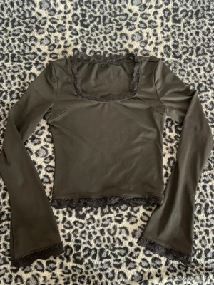 Mörkgrön långärmad topp med spets - Snygg mörkgrön långärmad topp med fyrkantig urringning och spetsdetaljer vid hals, ärmslut och nederkant. Croppad modell som passar perfekt till höga jeans eller kjol. Mjuk och stretchig känsla.