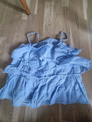 Ljusblå volangtop från Gina Tricot - Supersöt ljusblå topp från Gina Tricot med tunna axelband och flera volanger. Toppen är luftig och har en avslappnad passform. Aldrig använd men tagit av prislappen