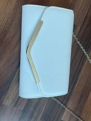 Vit kuvertväska med silverdetalj - Stilren vit kuvertväska med en snygg silverfärgad metallist på locket och avtagbar kedjerem i metall. Väskan har en mjuk, glittrig yta och en modern, kantig form. Perfekt för dig som vill ha en enkel men elegant accessoar till din outfit eller till skolbalen. (Pris går att diskutera) aldrig använd. 