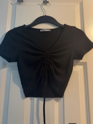 Svart ribbad croppad topp med snörning - Svart croppad topp från newyorker med ribbad struktur och snörning framtill. Toppen har v-ringning och korta ärmar. 