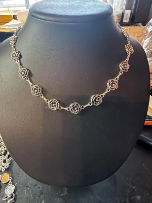 Silverfärgat halsband med blommor - Unikt halsband i silverfärgad metall med detaljerade blommor som länkas ihop runt hela halsen. Blommorna har en snygg retrovibe och ger halsbandet en cool och personlig touch. Perfekt för dig som gillar smycken med lite edge och fina detaljer.