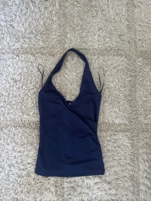 Mörkblå omlott halterneck topp - Snygg mörkblå halterneck topp med omlottdesign och djup v-ringning. Toppen är ärmlös och har tunna axelband perfekt sommartopp. Är i storlek xxs men jag brukar ha xs-s