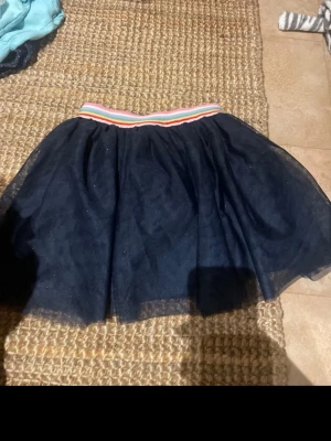 Svart tyllkjol med randig midja - BARN storlek Säljer en svart kortkjol i tyll med elastisk midja som har färgglada ränder i vitt, rosa och orange. Kjolen har en voluminös look och passar perfekt för dig som vill ha en lekfull och trendig stil.