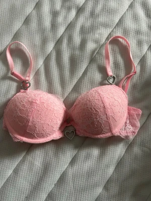 Rosa push-up bh med spets och hjärtan - Söt rosa push-up bh med vadderade kupor och spetsdetaljer. Axelbanden har dekorativa hjärtan i metall med små stenar och ett hjärta mellan kuporna. Bh:n har en romantisk och feminin look med fina spetskanter.