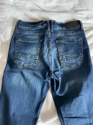 Blå raka jeans med kontrastsömmar - Säljer ett par klassiska blå jeans med raka ben och kontrastsömmar. Jeansen har två bakfickor och en svart patch vid linningen. Materialet är slitstarkt denim i bomull och passformen är normal. Perfekta till en avslappnad stil.