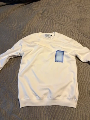 Vit sweatshirt från Casablanca - Vit sweatshirt från Casablanca med blått grafiskt tryck på bröstet och ryggen där det står 'Casa Blanca'. Tröjan har rund halsringning, långa ärmar och ribbade muddar. Perfekt för en clean och modern stil.