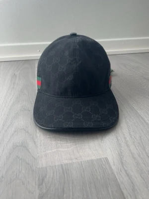 Svart keps med GG-mönster - Snygg svart keps med klassiskt GG-monogram över hela kepsen. På sidorna finns gröna och röda ränder som ger en ikonisk touch. Kepsen har böjd skärm och justerbar rem bak. Tillverkad i slitstarkt tyg som känns lyxigt.