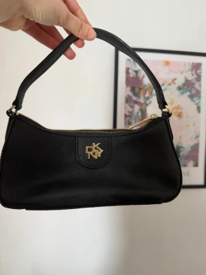 Svart axelremsväska från DKNY - Snygg svart handväska från DKNY med guldfärgade detaljer och dragkedja. Väskan har en stilren, avlång form och är tillverkad i ett strukturerat konstläder. Perfekt för dig som gillar klassisk och trendig design med en touch av lyx.