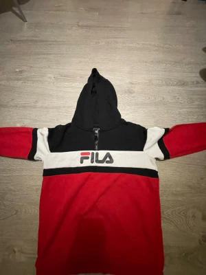 FILA hoodie i rött, svart och vitt - Snygg hoodie från FILA i färgerna rött, svart och vitt. Tröjan har en stor huva, halv dragkedja framtill och FILA-logga broderad på bröstet. Tillverkad i mjukt material som passar perfekt för en avslappnad stil.