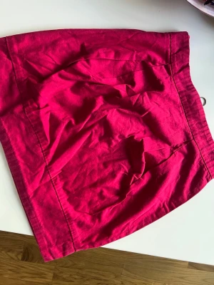 Rosa kortkjol i bomull - Snygg rosa kortkjol i mjukt bomullstyg. Kjolen har en enkel design med markerade sömmar och en liten metallring i midjan som detalj. Perfekt för att addera färg till din outfit.