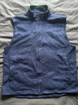 Mörkblå väst från Polo Ralph Lauren - Stilren mörkblå väst från Polo Ralph Lauren med dragkedja framtill, hög krage och diskret broderad logga på bröstet. Västen har fickor med dragkedja och är tillverkad i ett mjukt, mockaliknande material. Perfekt för lager på lager och en clean look.