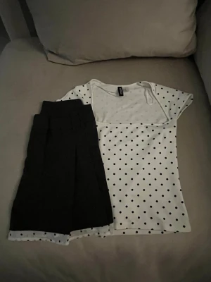 Prickigt topp med kortkjol med prickig kant- Set - Säljer en svart kortkjol med plisserad design och bred linning. Kjolen har en vit kant längst ner med svarta prickar som ger en snygg kontrast. Perfekt för dig som gillar en klassisk men lekfull stil. Märket är H&M.