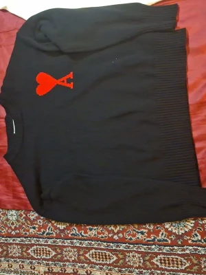 Svart stickad tröja från Ami - Svart stickad tröja från Ami med röd hjärtlogga på bröstet. Tröjan har rund halsringning och ribbade muddar vid ärmslut och nederkant. Perfekt för dig som gillar stilrena plagg med en twist. Storlek L
