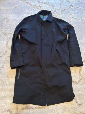 Svart rock från Lacoste - Stilren svart rock från Lacoste med klassisk krage och diskret logga på bröstet. Lång modell med raka linjer och dolda fickor framtill. Storlek M, Nypris 4500kr