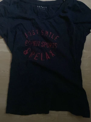Svart t-shirt från Esprit Sports - Cool svart t-shirt från Esprit Sports med röd text 'Just Smile & Relax' på bröstet. T-shirten har korta ärmar och en klassisk rund halsringning. Perfekt för dig som gillar en avslappnad och sportig stil.