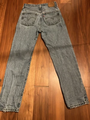 Levi's 501 ljusblå raka jeans - Klassiska Levi's 501 jeans i ljusblå tvätt med raka ben och hög midja. Jeansen har två bakfickor med den ikoniska Levi's-sömmen och läderpatch bak i midjan. Tillverkade i slitstark bomull, perfekt för en avslappnad och tidlös look. Har ett litet hål men syns nästan inte. Därför är priset så lågt. (Se på bilden) W27 L 29  Tveka inte på att ställa frågor! Priset är ej hugget i stenen. 🤩