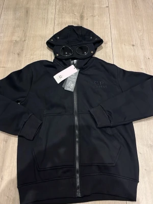 Svart hoodie från C.P. Company - Säljer en svart hoodie från C.P. Company med dragkedja framtill och två stora fickor. Huvan har inbyggda glasögon/goggles och loggan är broderad på bröstet. Tillverkad i mjukt material som känns skönt mot huden. Perfekt för dig som gillar streetwear och unika detaljer.
