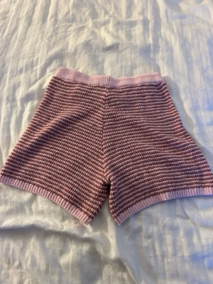 Rosa stickade shorts med snörning - Mysiga stickade shorts i ljusrosa och vitt randigt mönster. Shortsen har hög midja och snörning i midjan för justerbar passform. Perfekta för en avslappnad och trendig look.