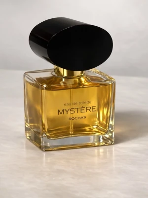Vintage Mystère Eau de Toilette från Rochas 50 ml - Mystère Eau de Toilette från Rochas i en elegant, fyrkantig glasflaska med gyllene vätska och en stor, svart oval kork. Flaskan har en lyxig känsla och modern design som sticker ut på parfymhyllan.