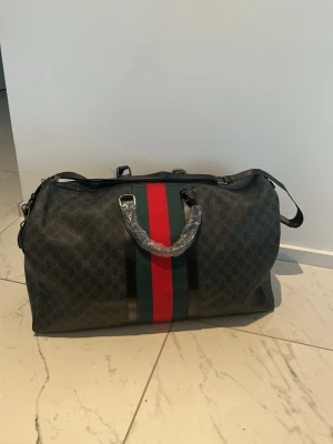 Gucci weekendbag med röd/grön rand - Snygg weekendbag från Gucci i mörkgrått med klassiskt GG-mönster. Väskan har breda ränder i rött och grönt, dubbla handtag och avtagbar axelrem. Perfekt för resor när du vill ha stil och funktion. Tillverkad i slitstarkt canvasmaterial med läderdetaljer.