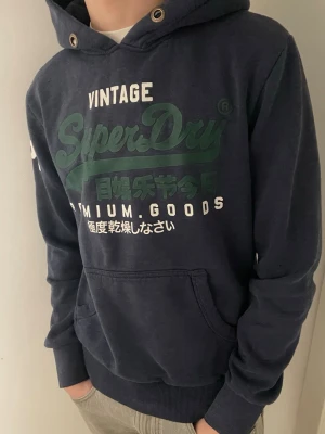 Mörkblå Superdry hoodie med tryck - Säljer en mörkblå hoodie från Superdry med grönt och vitt tryck på bröstet och ärmen. Tröjan har huva med snörning, känguruficka och ribbade muddar. Perfekt för chill dagar och streetstyle. Materialet är mjuk bomull och polyesterblandning. 