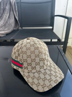 Beige GG keps från Gucci - Säljer en beige keps från Gucci med det klassiska GG-monogrammet över hela kepsen. På sidan finns en grön och röd rand som signaturdetalj. Kepsen har böjd skärm och justerbar rem bak. Materialet är canvas med exklusiv känsla.