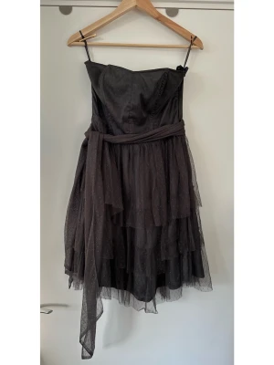 Svart grå axelbandslös H&M klänning i tyll, storlek 40 - Superfin axelbandslös grå klänning / festklänning / cocktailklänning från H&M Divided i storlek 40. Smickrande figurnära överdel med korsett-liknande inre band som håller toppen på plats. Härlig vid kjol i flera lager av tyll / mesh med små formvävda prickar i materialet. Klänningen har ett brett band i midjan som kan knytas framtill eller baktill. Tyllkjolen ger volym och en lekfull känsla. Superhärligt klänning i mycket fint skick. Älskar den, men tyvärr för liten för mig nu. 
