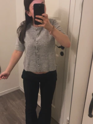 Grå kortärmad kofta med pärlknappar - Söt grå kofta med korta ärmar och rund hals. Koftan har pärlknappar framtill och är stickad i ett mjukt material. Passar perfekt till både jeans och kjol för en trendig look.