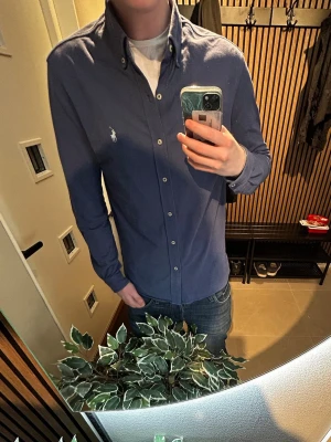 Blå skjorta från Ralph Lauren - Klassisk blå skjorta från Ralph Lauren med button-down krage och vit broderad logga på bröstet. Skjortan har långa ärmar, knappar framtill och är tillverkad i mjuk bomull. Perfekt till jeans eller chinos för en clean och stilren look.